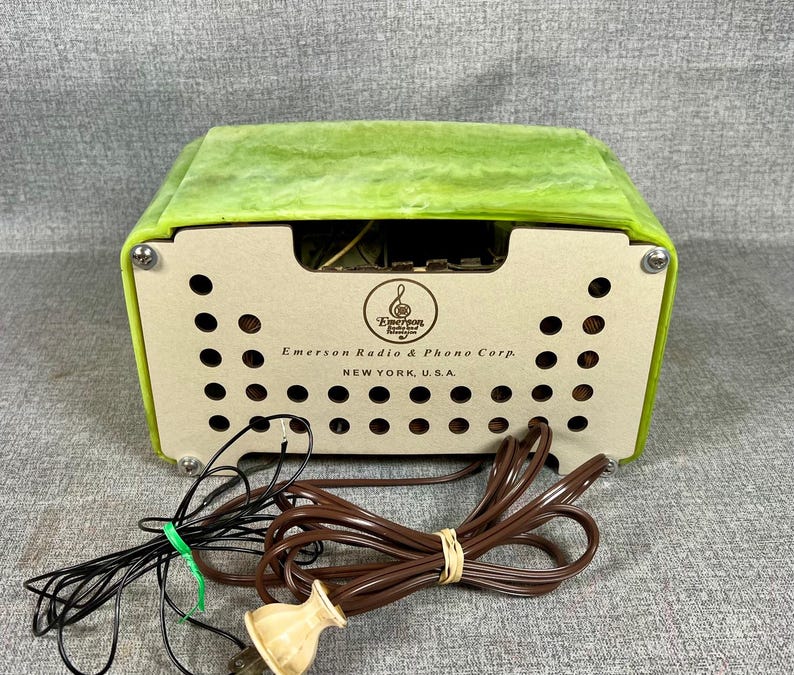 May include: A vintage Emerson Radio & Phono Corp. radio with a light green top and a beige front panel. The front panel has a series of circular holes and the company's logo and text. Black and brown cords with a plug are in the foreground.