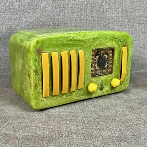 May include: A retro-style radio with a rectangular form, showcasing a marbled green finish and yellow details. The radio includes vertical yellow bars, a dial, and two yellow knobs. The dial has numbers and text.