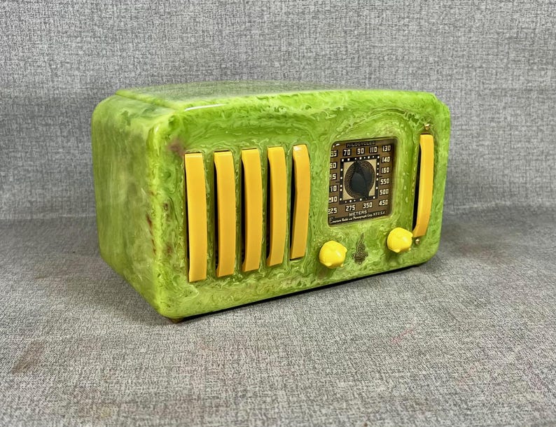 May include: A retro-style radio with a rectangular form, showcasing a marbled green finish and yellow details. The radio includes vertical yellow bars, a dial, and two yellow knobs. The dial has numbers and text.