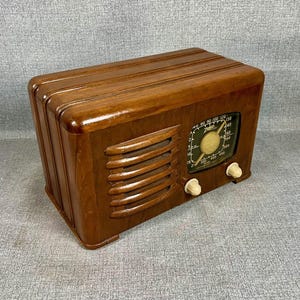 May include: A vintage wooden radio with a glossy finish. The radio features a speaker grill, a tuning dial, and two cream-colored knobs. The radio's face displays a green and white dial with the brand name "Zenith".