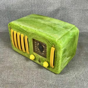 May include: A retro-style radio in a bright green colour with yellow detailing. The radio has a classic design with a dial, knobs, and speaker grill. The radio's face has a detailed display with numbers and a tuning dial. A decorative piece for home decor.