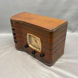 May include: A vintage wooden radio with a gold-colored dial and black knobs. The radio has a wood grain finish and is in good condition.