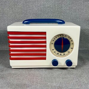 Pode incluir: Um r&aacute;dio de estilo vintage em uma caixa retangular branca com barras horizontais vermelhas no lado esquerdo. Possui uma al&ccedil;a azul na parte superior e um mostrador redondo com um ponteiro azul e vermelho. Dois bot&otilde;es azuis est&atilde;o abaixo.