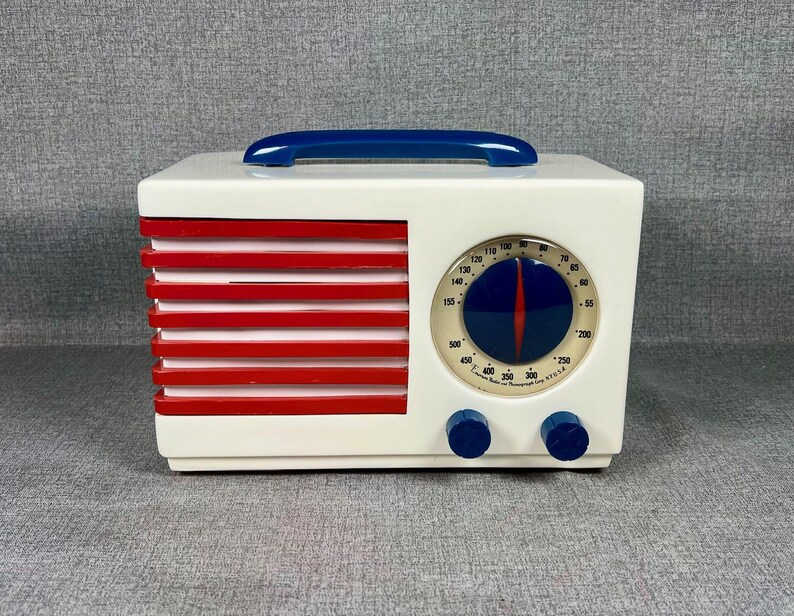 Pode incluir: Um r&aacute;dio de estilo vintage em uma caixa retangular branca com barras horizontais vermelhas no lado esquerdo. Possui uma al&ccedil;a azul na parte superior e um mostrador redondo com um ponteiro azul e vermelho. Dois bot&otilde;es azuis est&atilde;o abaixo.