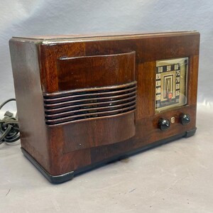 Może przedstawiać: Antyczne drewniane radio z ciemnobrązowym wykończeniem i srebrną płytą czołową. Radio ma tarczę z liczbami od 55 do 100 i pokrętło strojenia. Radio ma kratkę głośnika z przodu i dwa pokrętła z boku.