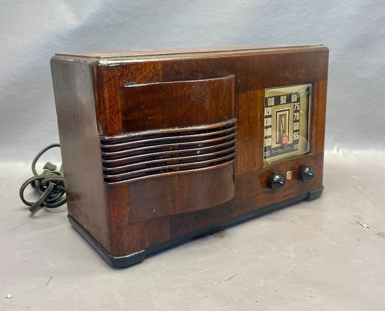 Może przedstawiać: Antyczne drewniane radio z ciemnobrązowym wykończeniem i srebrną płytą czołową. Radio ma tarczę z liczbami od 55 do 100 i pokrętło strojenia. Radio ma kratkę głośnika z przodu i dwa pokrętła z boku.