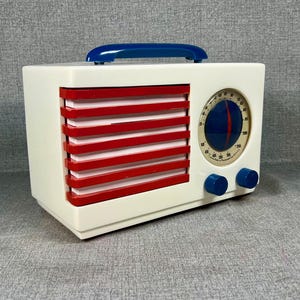 Pode incluir: Um r&aacute;dio de estilo vintage com corpo branco, ripas horizontais vermelhas e uma al&ccedil;a azul. O r&aacute;dio apresenta um mostrador circular com fundo azul e agulha vermelha, juntamente com dois bot&otilde;es azuis. O design lembra a est&eacute;tica de meados do s&eacute;culo.
