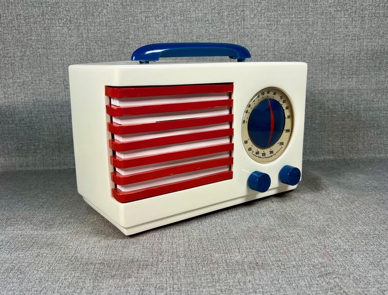 Pode incluir: Um r&aacute;dio de estilo vintage com corpo branco, ripas horizontais vermelhas e uma al&ccedil;a azul. O r&aacute;dio apresenta um mostrador circular com fundo azul e agulha vermelha, juntamente com dois bot&otilde;es azuis. O design lembra a est&eacute;tica de meados do s&eacute;culo.