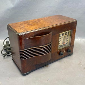 Może przedstawiać: Antena radiowa vintage wykonana z drewna, w ciemnobrązowym kolorze, z srebrnym pokrętłem. Pokrętło ma liczby od 55 do 100 i czerwoną wskaz&oacute;wkę. Radio ma dwa pokrętła z przodu.
