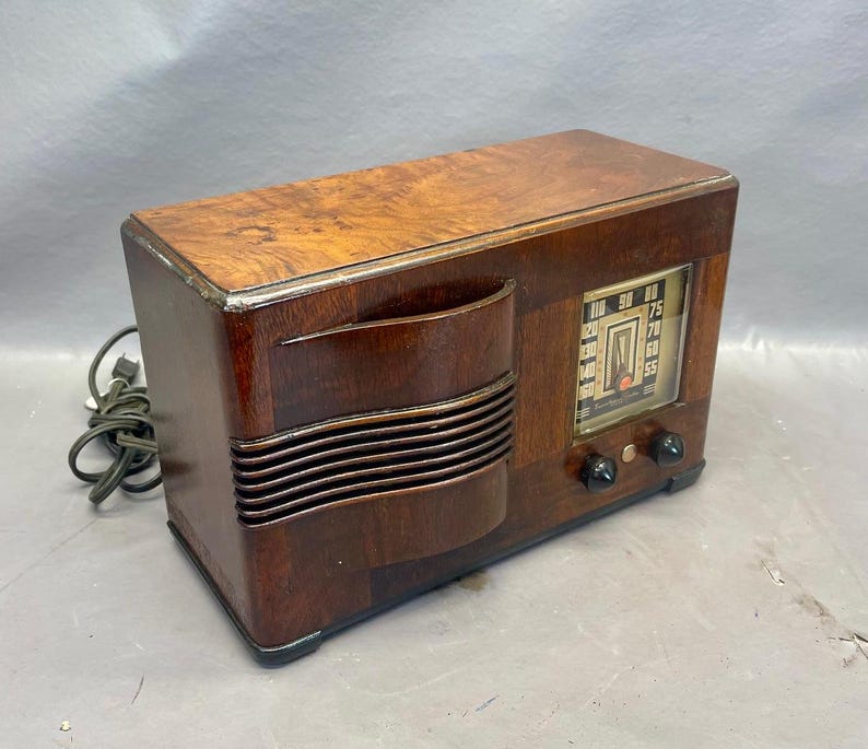 Może przedstawiać: Antena radiowa vintage wykonana z drewna, w ciemnobrązowym kolorze, z srebrnym pokrętłem. Pokrętło ma liczby od 55 do 100 i czerwoną wskaz&oacute;wkę. Radio ma dwa pokrętła z przodu.