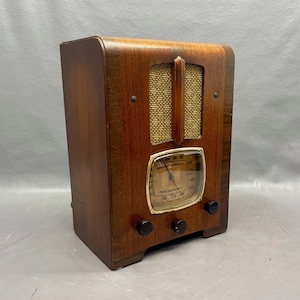 May include: A vintage wooden radio with a gold-colored dial and knobs. The radio has a woodgrain finish and a metal grille. The dial reads "BROADCAST & MUSIC" and "PUBLIC ADDRESS SYSTEMS".
