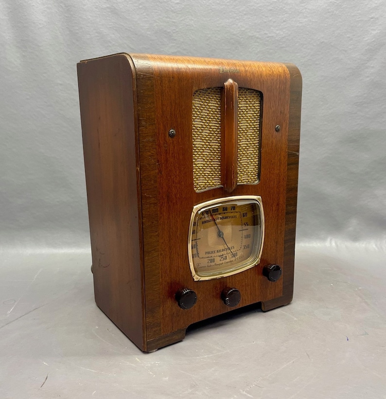 May include: A vintage wooden radio with a gold-colored dial and knobs. The radio has a woodgrain finish and a metal grille. The dial reads "BROADCAST & MUSIC" and "PUBLIC ADDRESS SYSTEMS".