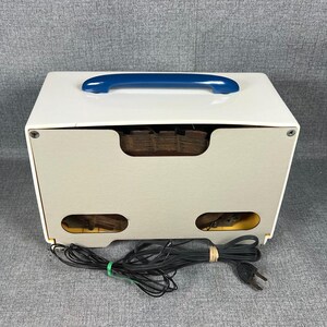 Pode incluir: Um r&aacute;dio vintage branco e bege com uma al&ccedil;a azul. O r&aacute;dio tem formato retangular com cantos arredondados e uma s&eacute;rie de pequenas aberturas na frente. Um cabo de alimenta&ccedil;&atilde;o preto est&aacute; conectado ao r&aacute;dio.