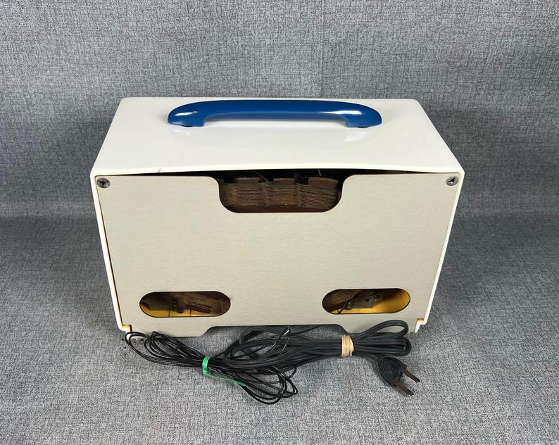 Pode incluir: Um r&aacute;dio vintage branco e bege com uma al&ccedil;a azul. O r&aacute;dio tem formato retangular com cantos arredondados e uma s&eacute;rie de pequenas aberturas na frente. Um cabo de alimenta&ccedil;&atilde;o preto est&aacute; conectado ao r&aacute;dio.