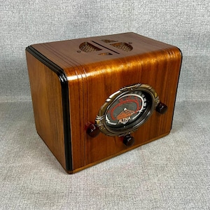 May include: A wooden radio with a polished brown finish and black trim. The front features a circular brass-toned dial with the text AETNA, a red frequency scale, and three dark brown control knobs. The top has two decorative speaker grilles.
