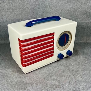 Pode incluir: Um r&aacute;dio de estilo vintage em uma caixa retangular branca com ripas horizontais vermelhas e uma al&ccedil;a azul. O r&aacute;dio apresenta um mostrador circular com um design azul e vermelho e dois bot&otilde;es azuis.