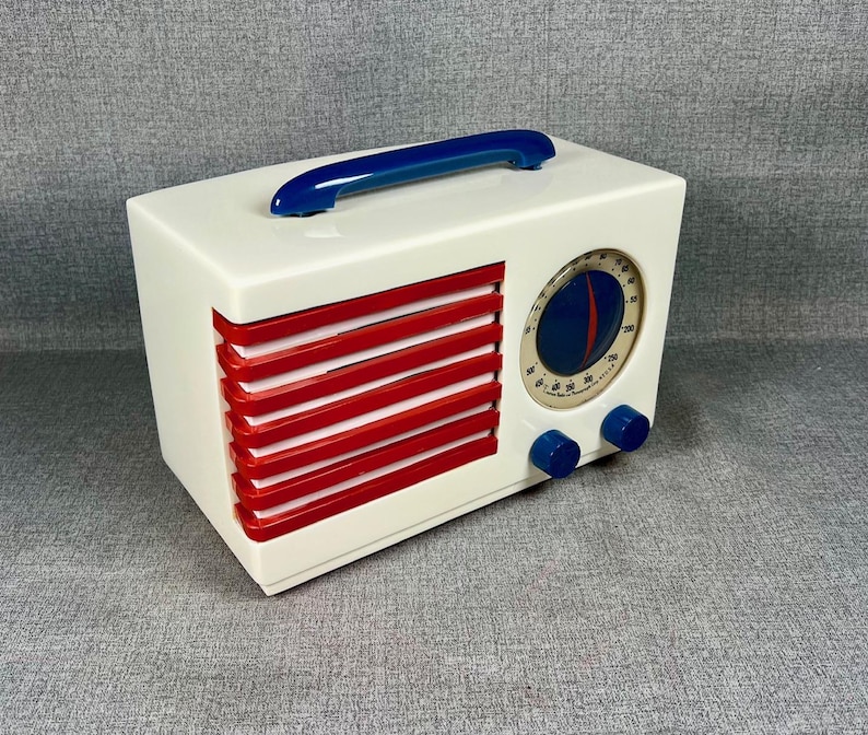 Pode incluir: Um r&aacute;dio de estilo vintage em uma caixa retangular branca com ripas horizontais vermelhas e uma al&ccedil;a azul. O r&aacute;dio apresenta um mostrador circular com um design azul e vermelho e dois bot&otilde;es azuis.