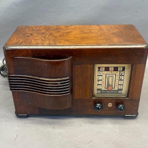 Może przedstawiać: Antyczne drewniane radio z brązową drewnianą obudową i srebrną płytą czołową. Radio ma pokrętło z liczbami od 55 do 160 i tekstem "Frequency Range".
