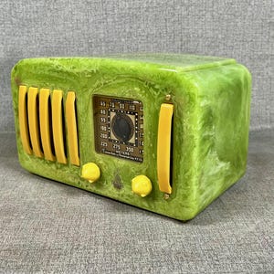May include: A vintage green radio with yellow detailing. The radio has a rectangular form with a dial and speaker grilles. The face of the radio features a dial and a small display with numbers. The radio is a retro style.