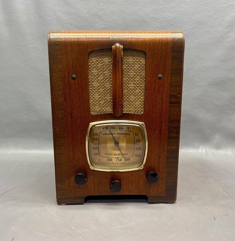 May include: A vintage wooden Emerson radio with a gold-colored dial and a speaker grille. The dial has markings for "Broadcast Band" and "Police Kilocycles".