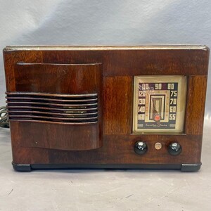 Może przedstawiać: Antyczne drewniane radio z ciemnobrązowym wykończeniem i srebrnym pokrętłem. Pokrętło ma czarną wskaz&oacute;wkę i białe cyfry. Radio ma dwa pokrętła z przodu.