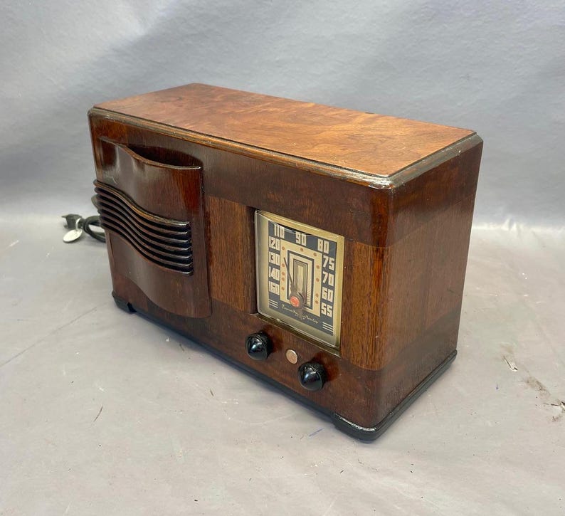 Może przedstawiać: Antyczne drewniane radio z brązową drewnianą obudową i srebrną płytą czołową. Radio ma tarczę z liczbami od 55 do 110. Radio ma dwa pokrętła z przodu.
