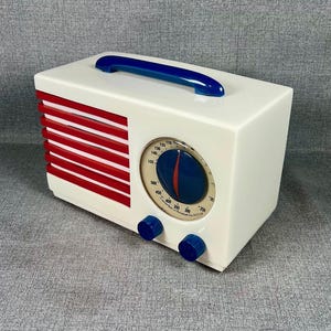 Pode incluir: Um r&aacute;dio de estilo retr&ocirc; em uma caixa retangular branca com uma al&ccedil;a azul. O r&aacute;dio apresenta uma grelha de alto-falante vermelha e branca, um mostrador circular e dois bot&otilde;es azuis. O mostrador tem um ponteiro azul e vermelho.
