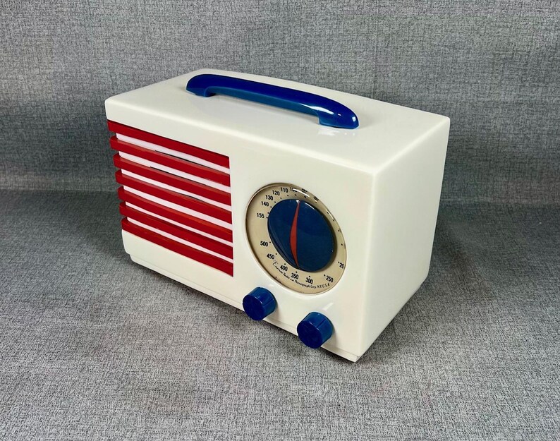Pode incluir: Um r&aacute;dio de estilo retr&ocirc; em uma caixa retangular branca com uma al&ccedil;a azul. O r&aacute;dio apresenta uma grelha de alto-falante vermelha e branca, um mostrador circular e dois bot&otilde;es azuis. O mostrador tem um ponteiro azul e vermelho.