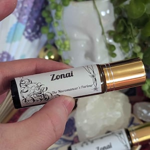 May include: A brown glass essential oil roller bottle with a gold cap. The label reads "Zonai" and "The Necromancer's Parlour" with decorative black line art. Another bottle is visible in the background.