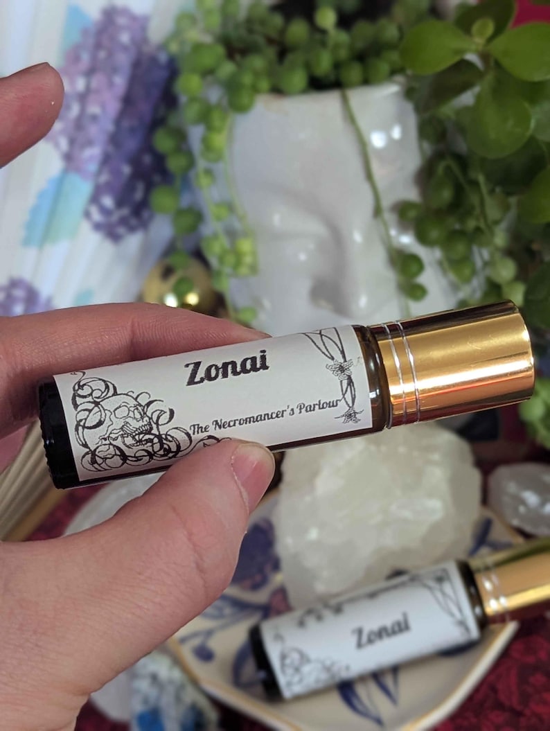 May include: A brown glass essential oil roller bottle with a gold cap. The label reads "Zonai" and "The Necromancer's Parlour" with decorative black line art. Another bottle is visible in the background.