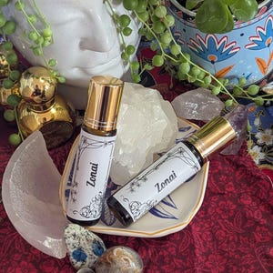 May include: Two roll-on perfume bottles with gold caps and white labels reading "Zonai" are displayed on a small decorative plate. The bottles are surrounded by crystals and decorative items, including a white ceramic face and a small dish.