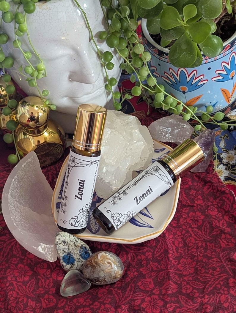 May include: Two roll-on perfume bottles with gold caps and white labels reading "Zonai" are displayed on a small decorative plate. The bottles are surrounded by crystals and decorative items, including a white ceramic face and a small dish.
