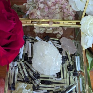 May include: A gold-framed glass box containing small perfume vials, crystals, and dried flowers. The vials have black caps and labels. A large, clear crystal and a pink rose are visible.