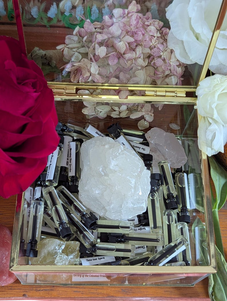 May include: A gold-framed glass box containing small perfume vials, crystals, and dried flowers. The vials have black caps and labels. A large, clear crystal and a pink rose are visible.