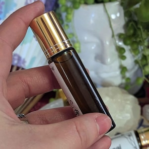 May include: A small, brown glass essential oil roller bottle with a gold cap. The bottle has a white label with text and decorative elements. The bottle is held in a hand, with other bottles visible in the background.