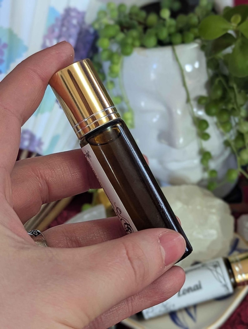 May include: A small, brown glass essential oil roller bottle with a gold cap. The bottle has a white label with text and decorative elements. The bottle is held in a hand, with other bottles visible in the background.