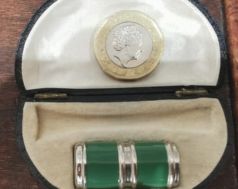 May include: A green and silver cylindrical object in a black velvet lined box. The box is open and a British one pound coin is visible in the top of the box.