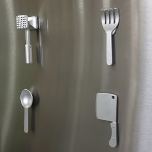 May include: Four silver kitchen utensil magnets, including a meat mallet, a fork, a spoon, and a butcher knife, are attached to a stainless steel refrigerator.