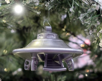 May include: A silver-colored, 3D-printed UFO Christmas ornament hanging on a green Christmas tree. The ornament has a classic flying saucer design with a circular body and detailed underside. The ornament is suspended by a small metal loop.