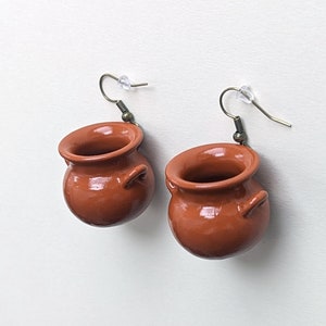 May include: A pair of terracotta pot earrings with brown clay pots and silver hooks. The pots have a rounded shape with a small opening at the top and two small handles on the sides.