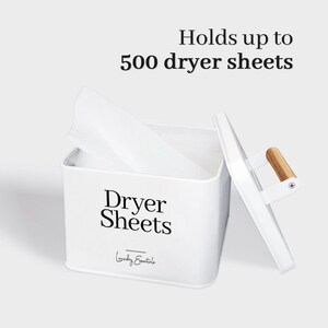 May include: White metal storage container with a wooden handle, labeled "Dryer Sheets" and "Laundry Essentials". The container is open and has a dryer sheet inside. The text "Holds up to 500 dryer sheets" is above the container.