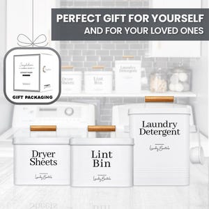 May include: Three white metal containers with wooden handles and black labels. The containers are labeled "Laundry Detergent", "Dryer Sheets", and "Lint Bin". The containers are on a white surface in front of a white tiled wall.