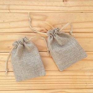 May include: Two small, beige burlap drawstring bags with white string ties. The bags are on a light brown wooden surface.