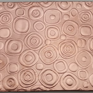 May include: A close-up of a copper sheet with a textured pattern of circles and ovals. The sheet has a warm, metallic sheen.