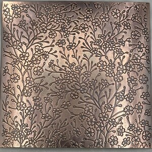 May include: Square copper-coloured metal plate with an embossed floral design. The pattern features delicate flowers and winding branches, creating a textured surface. The overall effect is a detailed, nature-inspired decorative element.