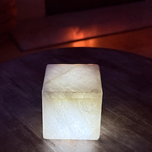 Puede incluir: Una caja cuadrada de alabastro brillante con tapa, sobre una superficie de madera oscura. La piedra translúcida tiene vetas sutiles y una luz suave y cálida. La caja es un objeto decorativo. Dimensiones: aproximadamente 10 cm x 10 cm x 10 cm.