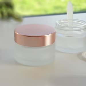 Könnte beinhalten: Zwei mattierte Glasbehälter mit roségoldenen Deckeln. Ein Behälter hat einen durchsichtigen Deckel und einen weißen Spatel im Inneren. Die Behälter stehen auf einer weißen Oberfläche mit einer braunen Nuss und einem grünen Blatt.