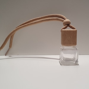 May include: A clear glass bottle with a wooden cap and a beige string for hanging. The bottle is square shaped and is small in size.