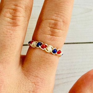 May include: A gold wire ring with red, white, and blue beads woven into the wire. The ring is on a finger.