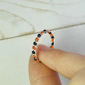 May include: A silver ring with a braided design, featuring alternating black and orange beads. The ring is held between fingers, showcasing its intricate details and color combination. The ring is a piece of jewelry.